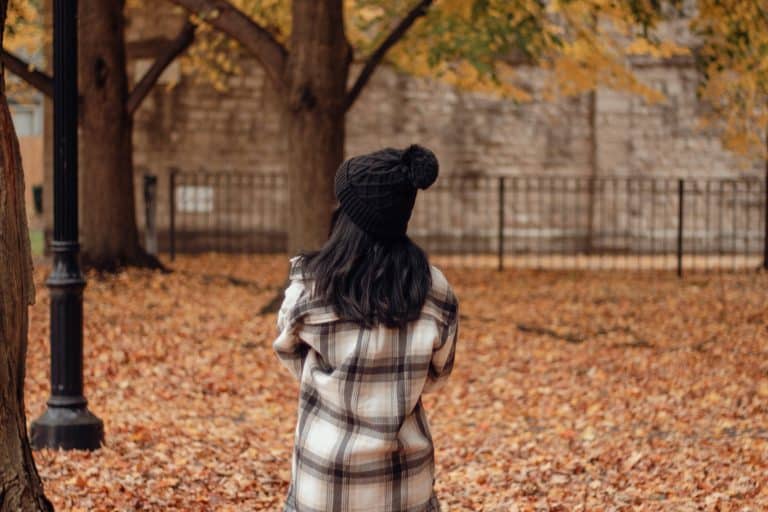 Woman wearing a black knit hat and plaid coat walking through a tree-lined park covered in autumn leaves.